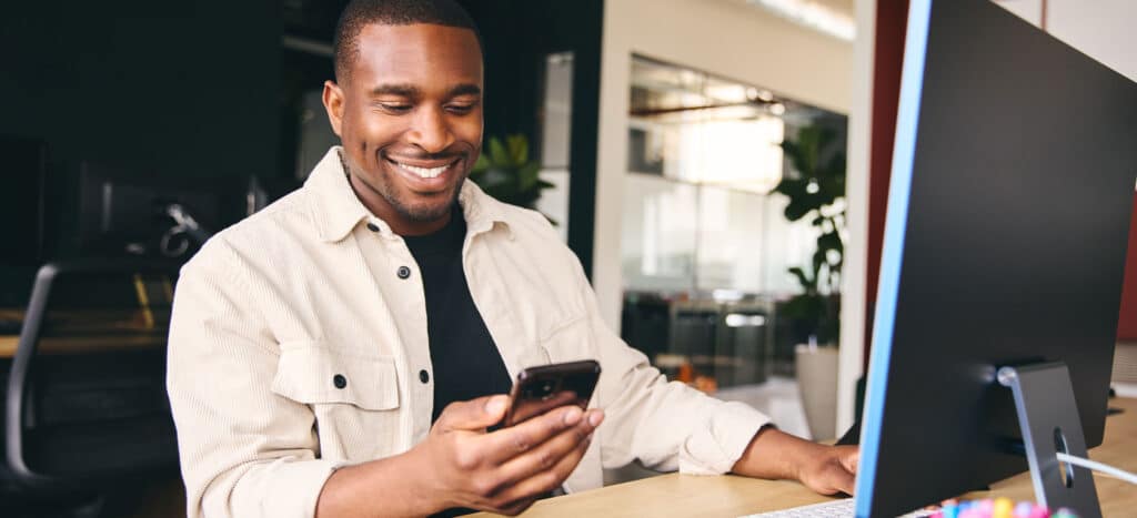 Man smiling at cell phone in office space sitting at a desk with computer