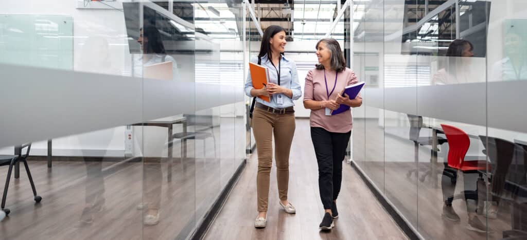 Two women walking through office space, carrying notebooks