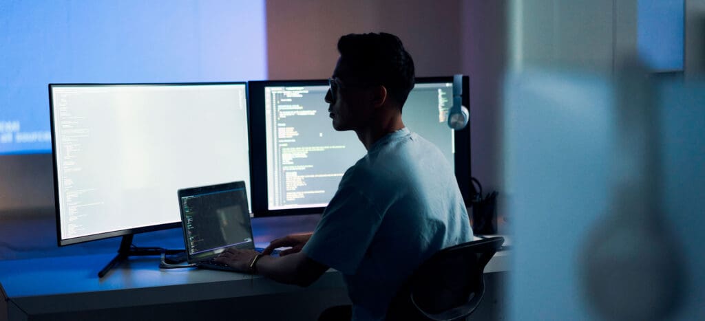 Software developer working on laptop, with two monitors displaying code behind it