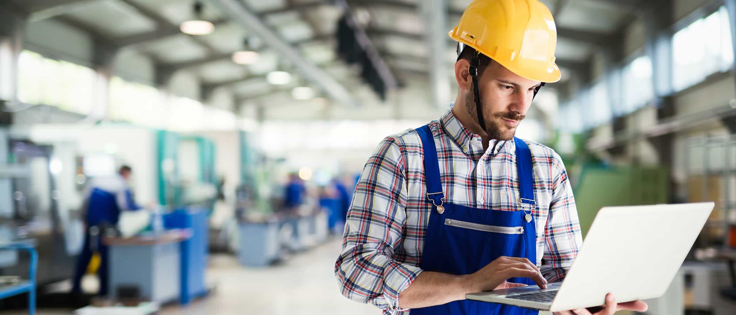 Man in manufacturing plant wearing a hardhat, typing on a laptop