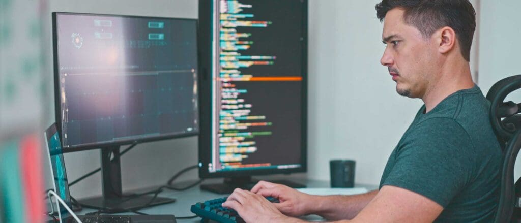 Man typing on keyboard with two monitors displaying code in front of him