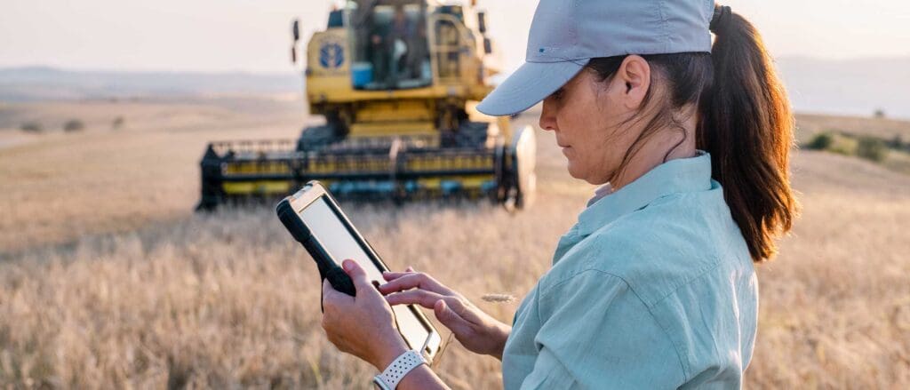 Woman in farm field with tractor behind her, typing on tablet