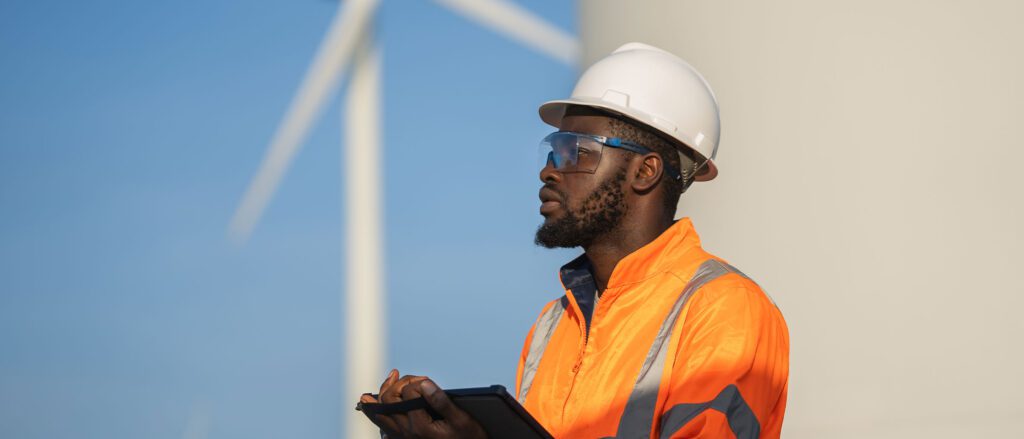 Man wearing a hardhat, holding a clipboard with windmills in the background