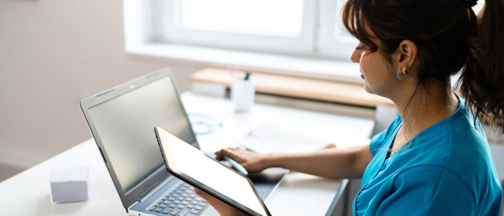 Medical professional reading results from tablet and entering data on a laptop